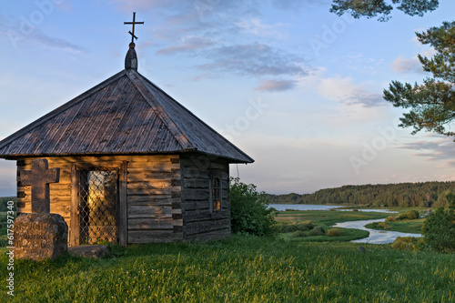 Landscape with an old wooden chapel at sunset against a background of a green meadow and a river