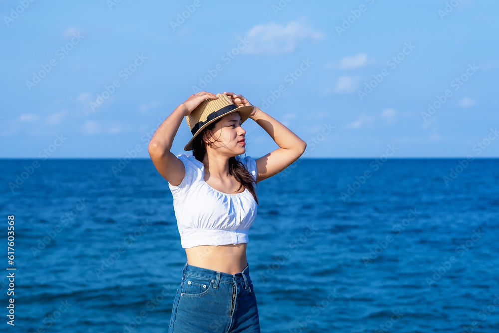 Lifestyle image of carefree traveling woman standing on sandy beach near turquoise colored ocean in summertime. Female tourist with straw hat in hands enjoying traveling to exotic nature leisure time.