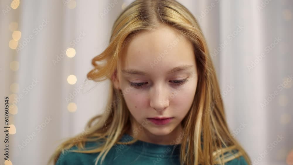 Portrait of pleasant blonde blue eyed teen girl at home touching hair looking at camera and down. Teenager preening, wearing emerald sweatshirt. Everyday usual life, sincere human emotion concept. Portrait of pleasant blonde blue eyed teen girl at home touching hair looking at camera and down. Teenager preening, wearing emerald sweatshirt. Everyday usual life, sincere human emotion concept.