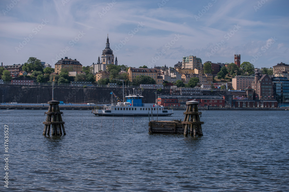An inner harbor ferry passing anchor buoys, the cliff Stadsgården in the district Södermalm, old apartment houses and the church Katarina kyrka, a sunny summer morning in Stockholm