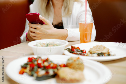 Young woman freelancer sitting at table with dishes in cafe and holding phone. Concentrated lady working at lunch time.