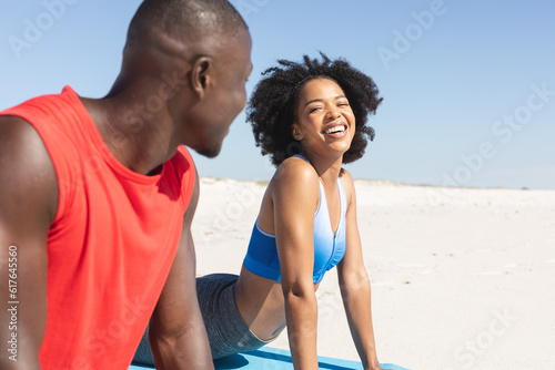 Happy, fit african american couple practicing yoga, stretching and smiling on sunny beach