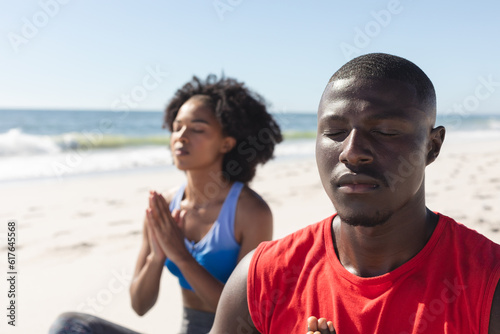 Happy, fit african american couple practicing yoga meditation sitting on sunny beach
