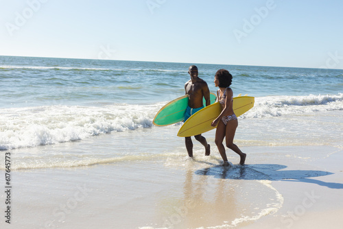 Happy, fit african american couple carrying surfboards walking on sunny beach in the sea, copy space