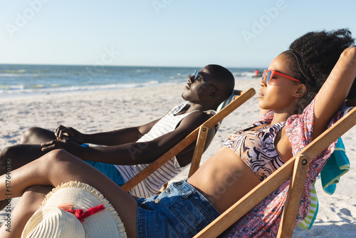 Happy african american couple in sunglasses sitting in deckchairs relaxing on sunny beach by the sea © wavebreak3