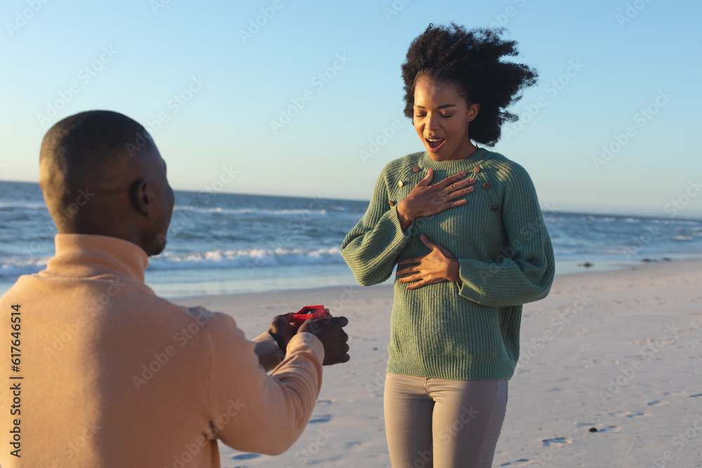 © wavebreak3 - Happy african american man kneeling with ring proposing to surprised girlfriend on sunny beach