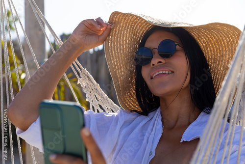 Obraz na plátne Happy biracial woman in sunglasses and sunhat sitting in hammock taking selfie o