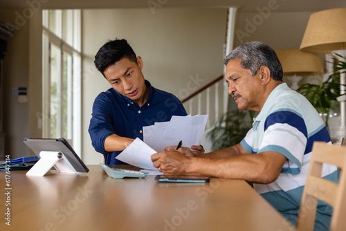Diverse financial advisor and senior man discussing paperwork and using tablet in dining room