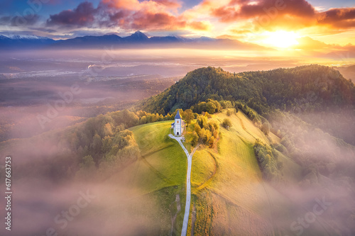 View from above of the serene beauty of the small Church of St. Primoz in Jamnik caressed by the fog at sunrise on green hill in Slovenia. Julian Alps, Kranj, Gorenjska