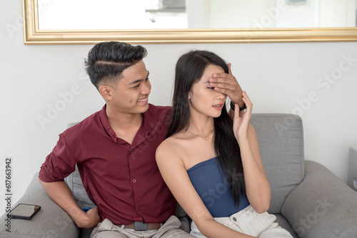 A young handsome man covers his girlfriend eyes while getting out a small gift behind his back. A playful scene at home.