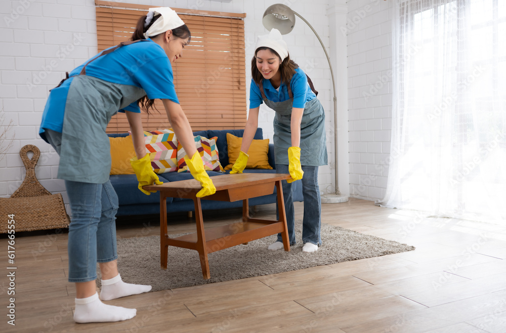 Two young women in uniform and gloves cleaning the living room at home, lifting the table Two young women in uniform and gloves cleaning the living room at home, lifting the table