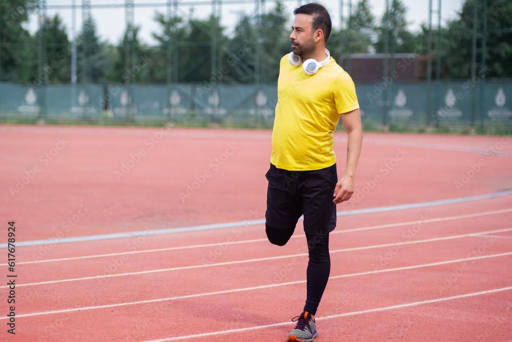 Young man with wireless headphones warming up leg muscles before training on running track with rubber surface and white lines at sports ground in city park