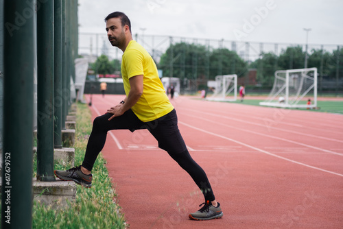 Athletic man in tracksuit doing lunges stretching legs leaning on fence support at urban stadium runner warming up training on sports arena with football field