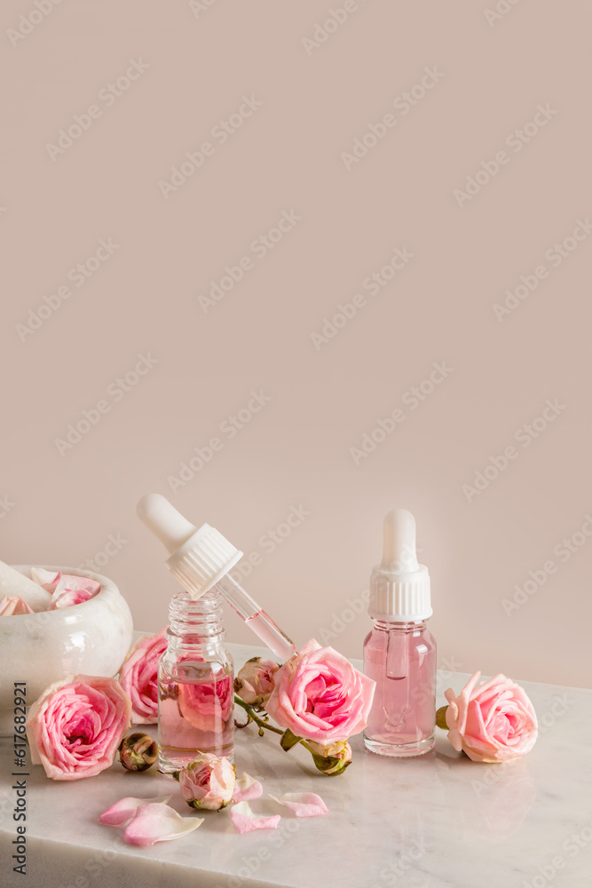 An open bottle and rose oil filled with a pipette, a bottle of cosmetic product among small roses on a beige background. Vertical view. A copy space. An open bottle and rose oil filled with a pipette, a bottle of cosmetic product among small roses on a beige background. Vertical view. A copy space.