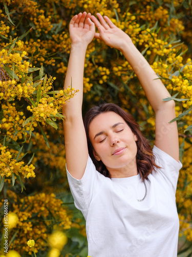 Beautiful young woman with dark curly hair and perfect skin wearing white tshirt posing near blooming flowers blooming mimosa. Nude make up. Allergy season, antihistamines. Aroma oils natural perfumes