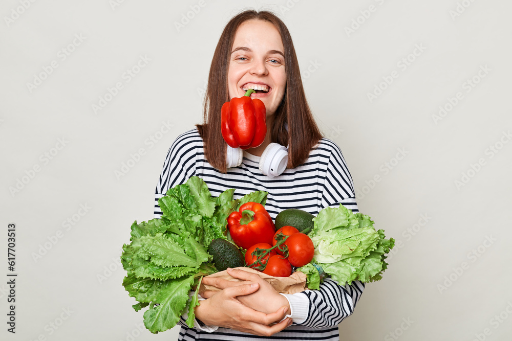 Funny laughing brown haired young woman embraces bouquet of fresh vegetables wearing striped casual shirt holding red pepper in mouth posing isolated over gray background Funny laughing brown haired young woman embraces bouquet of fresh vegetables wearing striped casual shirt holding red pepper in mouth posing isolated over gray background