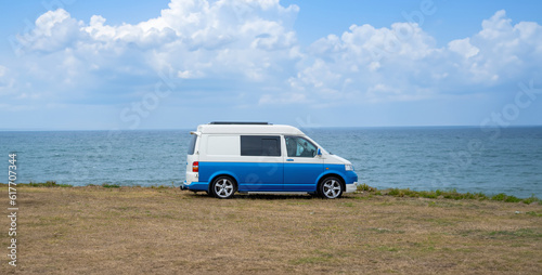 Blue vintage camper van parked on a grassy plain with the Atlantic Ocean in the background under a semi-cloudy sky lit by the early morning sun. © sirbouman