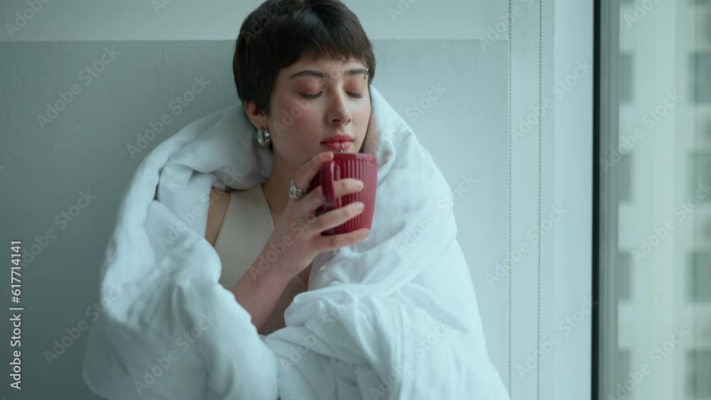 Depressed confident woman with piercings drinking tea and looking at view covered with blanket at home Depressed confident woman with piercings drinking tea and looking at view covered with blanket at home