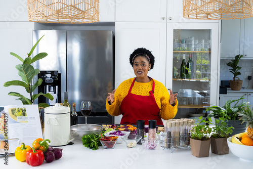 Happy plus size african american cooking vlogger in apron preparing meal and filming in kitchen