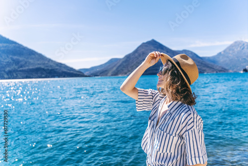 A woman in a hat standing on the seashore overlooking the mountains. Summer holidays and travel concept