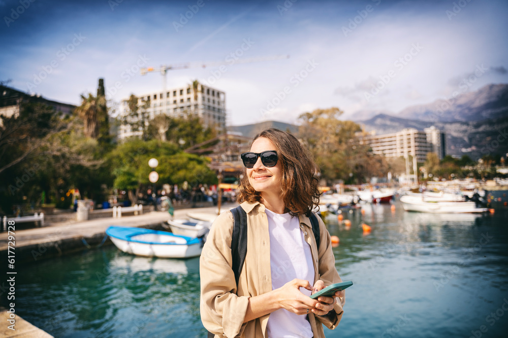 Young stylish happy woman in sunglasses standing on the sea promenade in the city with a smartphone
