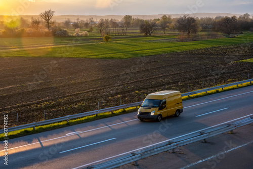 Yellow modern delivery small shipment cargo courier van moving fast on motorway road to city urban suburb. Beautiful sunset in the background. 