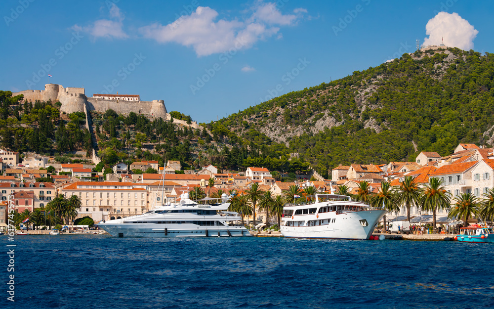 Panoramic view of croatian holiday hot spot old village of Hvar on Hvar island in the Adriatic Sea. Waterfront promenade with motor yachts and fisher boats. Fortica fortress on the hills above town.