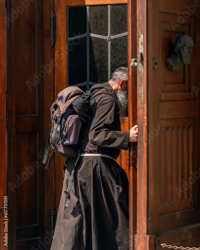 Authentic Catholic pilgrim in black clothes with a large backpack behind his back, girded with a white rope, opens a large wooden door, close-up of a monk in the sunlight at the entrance to the temple photo