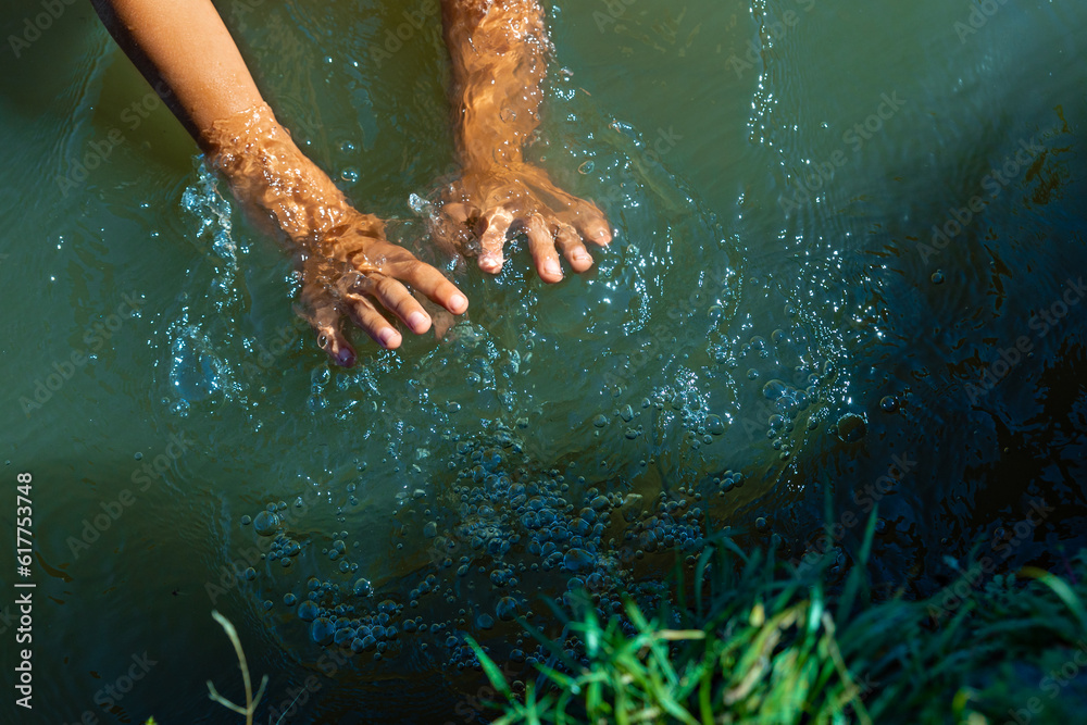 Fototapeta premium The child's hands spray dense green water in the lake. Summer activity for children - a game with clear water