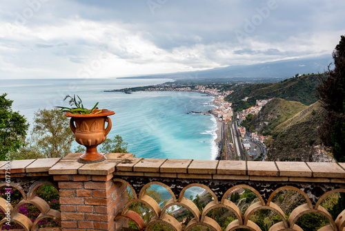 Mediterranean flowering plant in a stone ceramic pot with scenic view on seaside and Mount Etna volcano from public garden Parco Duca di Cesaro to Giardini Naxos in Taormina, Sicily, Italy