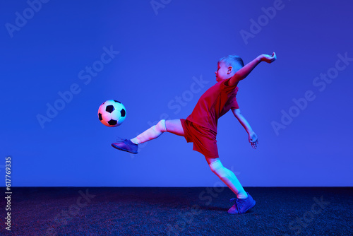 One sportive child, boy in red uniform playing, training football over dark blue background in neon. Concept of action, sportive lifestyle, team game, health, energy