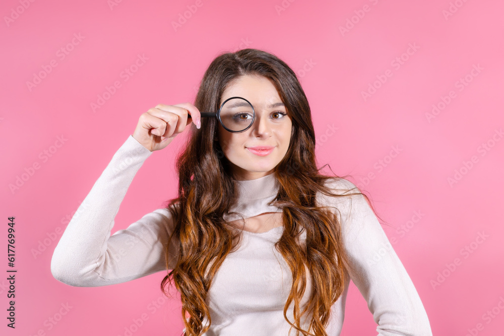 Young beautiful woman holding a magnifying glass near her eye on the pink background. Young beautiful woman holding a magnifying glass near her eye on the pink background.