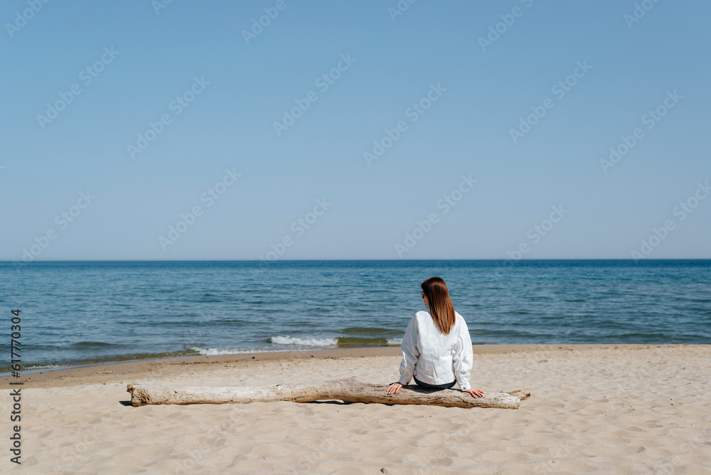 Lonely young woman in a white jacket sitting alone on a log on beach by sea, mood. Rear view of a woman in casual clothes alone on shore, copy space