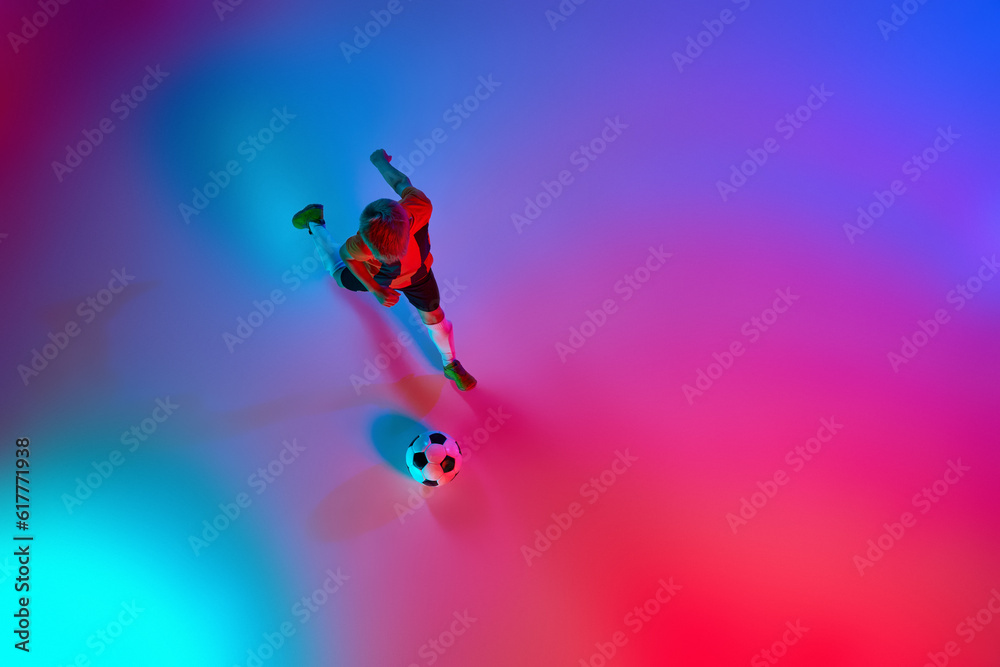 Obraz premium Aerial view of playing little boy during training with football ball in gradient red-blue neon light. Concept of action, sportive lifestyle, team game, health, energy