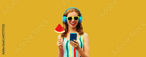 Summer portrait of happy smiling young woman model in headphones listening to music on smartphone with juicy lollipop or ice cream shaped slice of watermelon on yellow background