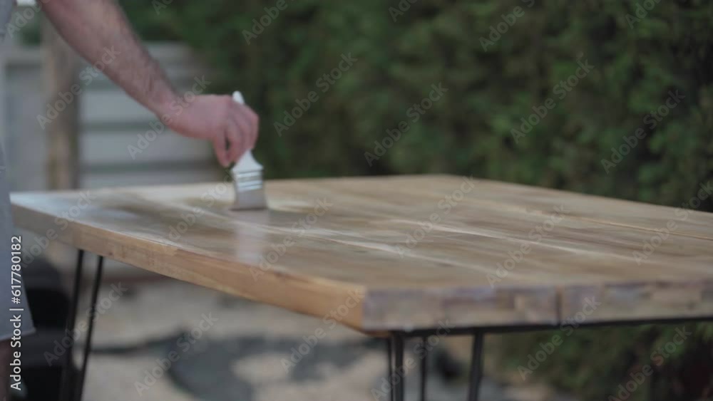 Wooden table on backyard with unrecognizable Caucasian man painting furniture. Male carpenter on the left applying primer on wood with brush Wooden table on backyard with unrecognizable Caucasian man painting furniture. Male carpenter on the left applying primer on wood with brush