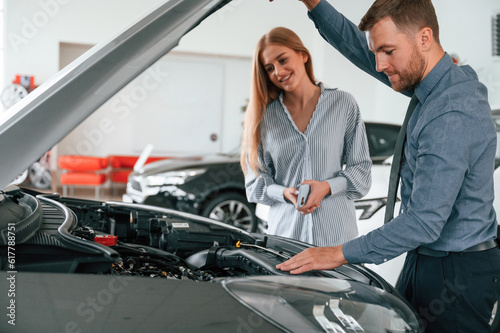 Showing engine under the hood. Man in formal clothes is with woman customer with the electric car