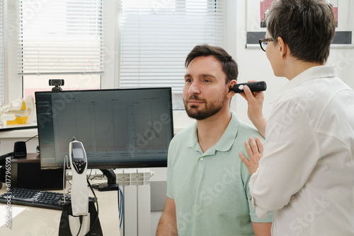 Man has appointment at clinic, checking hearing. Doctor audiologist consulting at hearing rehabilitation clinic to select digital device for middle age man. Doctor consulting patient photo