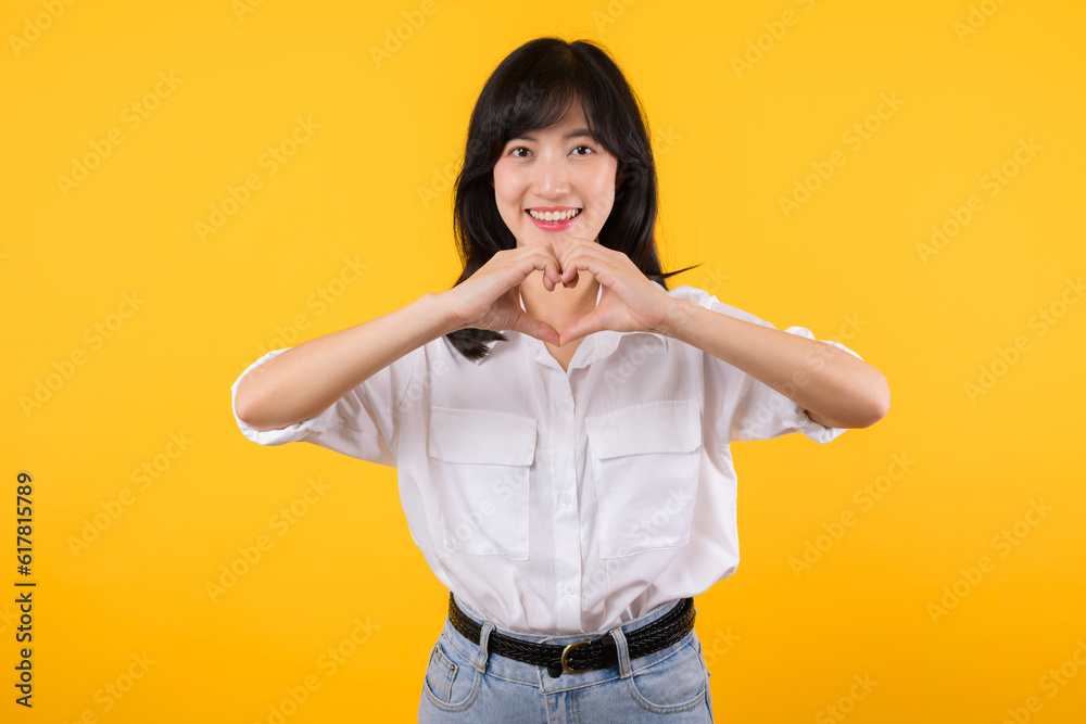 Young female healthcare. Asian woman wearing white shirt feels happy and romantic shapes heart gesture expresses tender feeling poses isolated on yellow background. People affection and care concept. Young female healthcare. Asian woman wearing white shirt feels happy and romantic shapes heart gesture expresses tender feeling poses isolated on yellow background. People affection and care concept.