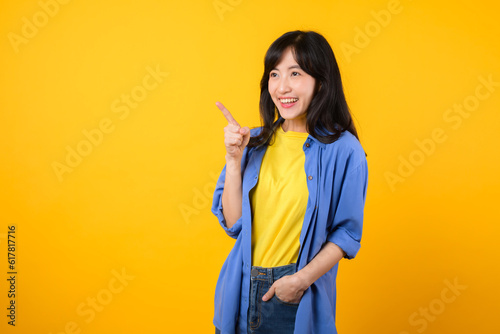Capture attention with engaging portrait. A young Asian woman wearing a yellow t-shirt and blue shirt showing happy smile while pointing finger to free copy space. education promotion concept.