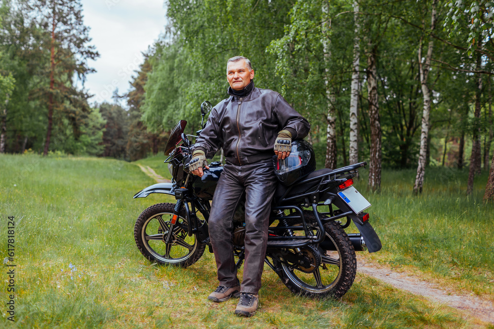 Fototapeta premium Elderly biker leaning on motobike in summer forest. Man in leather clothes relaxing enjoying nature.