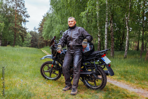 Elderly biker leaning on motobike in summer forest. Man in leather clothes relaxing enjoying nature.