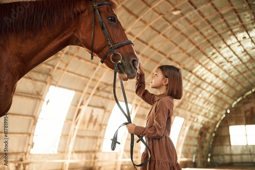 Side view. Cute little girl is with horse indoors