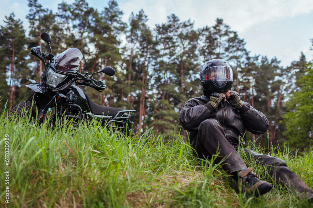 Close up of biker unfastening helmet after riding motobike in summer forest. Man wearing leather clothes outdoors. Close up of biker unfastening helmet after riding motobike in summer forest. Man wearing leather clothes outdoors.