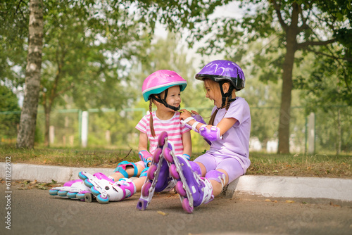 A girl helps her younger sister fasten a protective sports helmet for roller skating together in the suburbs. Children have fun doing their favorite sports hobby together