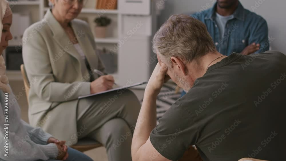 Back view of depressed mature soldier attending group therapy session with Asian female psychologist trying to help