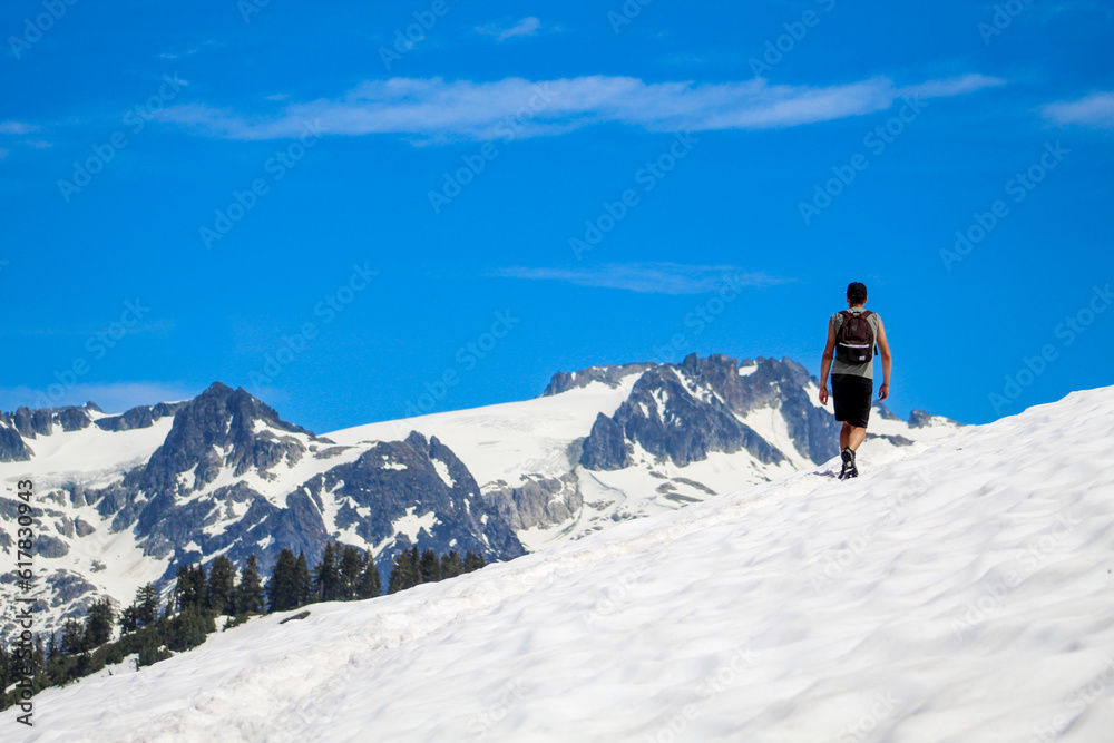 Man hiking alone on snow covered trail in summer with blue sky overlooking alpine mountains