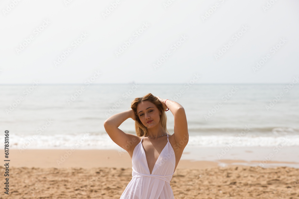 Young beautiful blonde woman in white dress is walking on the sand on the shore of the beach on a sunny day. The woman makes different body expressions. In the background the blue sea. Young beautiful blonde woman in white dress is walking on the sand on the shore of the beach on a sunny day. The woman makes different body expressions. In the background the blue sea.