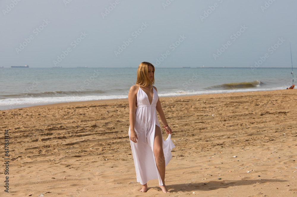 Young beautiful blonde woman in white dress is walking on the sand on the shore of the beach on a sunny day. The woman makes different body expressions. In the background the blue sea. Young beautiful blonde woman in white dress is walking on the sand on the shore of the beach on a sunny day. The woman makes different body expressions. In the background the blue sea.