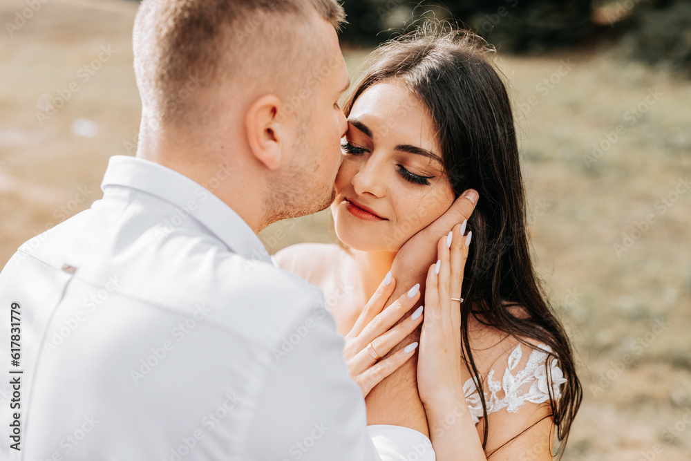 A stylish couple of European newlyweds. A smiling bride in a white dress hugs the groom from behind. The groom is dressed in a white shirt, without a tie. Outdoor wedding in the mountains A stylish couple of European newlyweds. A smiling bride in a white dress hugs the groom from behind. The groom is dressed in a white shirt, without a tie. Outdoor wedding in the mountains